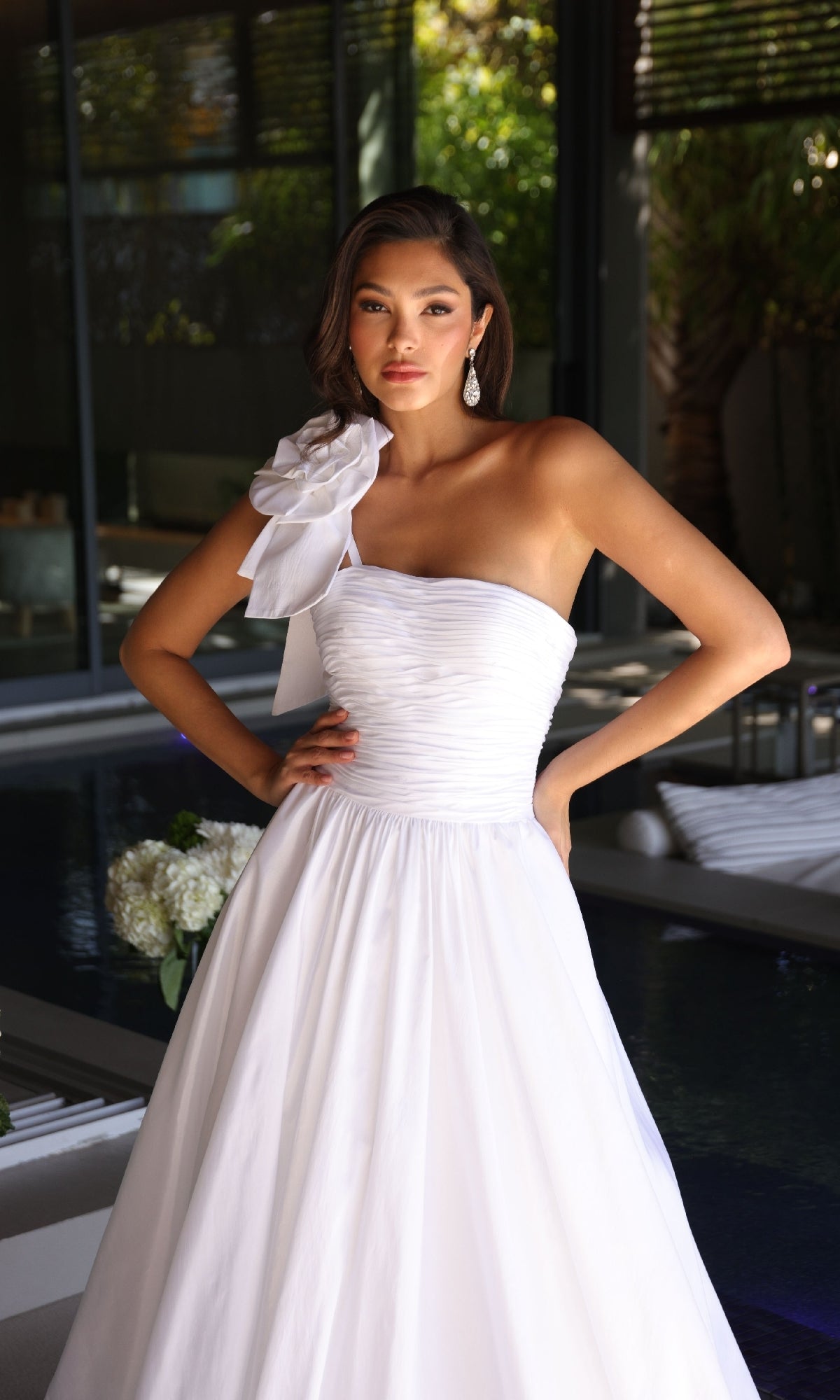 A woman stands indoors with hands on hips, wearing the Ava Presley 26524 long prom dress—a white, one-shoulder gown featuring a large shoulder bow and timeless elegance.